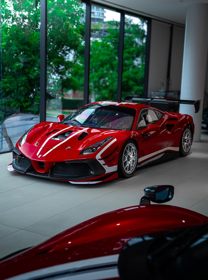 Red Ferrari 488 Challenge Evo with white racing stripes and a fixed rear wing, front three-quarter angle on a glass-fronted showroom floor.
