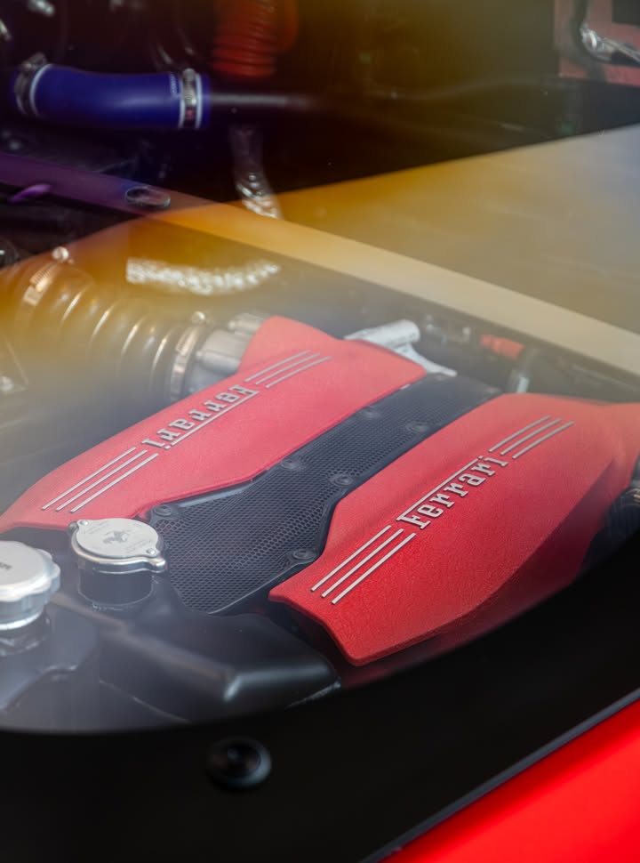Ferrari V8 engine bay seen through the glass hatch, red crackle-finish cam covers either side of a carbon intake plenum.