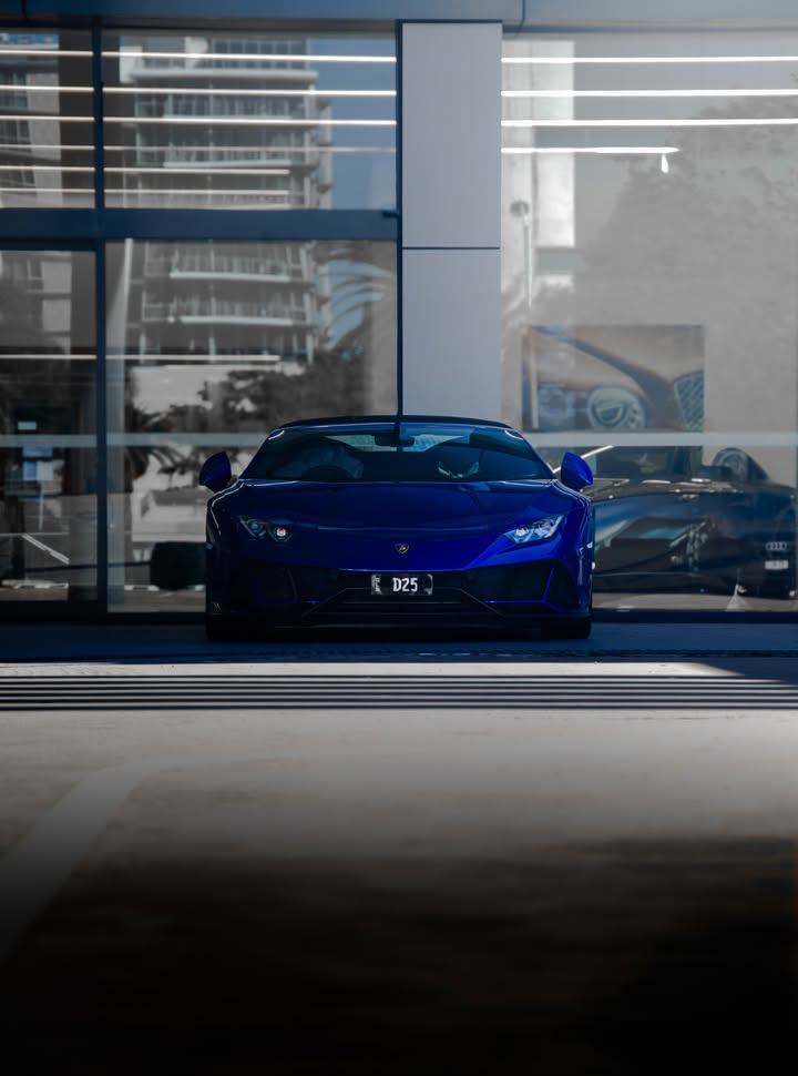 Blue Lamborghini Huracan EVO Spyder head-on in shadow, high-rise towers reflected in the dealership glass behind it.