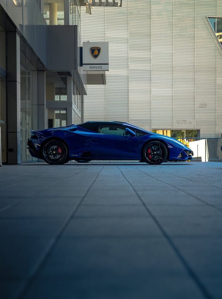 Blue Lamborghini Huracan EVO Spyder in profile beneath a Lamborghini service sign, shot low across the forecourt pavers.