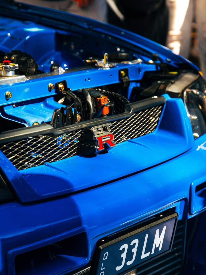 GT-R badge on the grille of a Bayside Blue Nissan Skyline R34 with the bonnet raised, Queensland plate below.