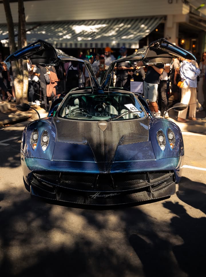 Blue exposed-carbon Pagani Huayra parked with both gullwing doors raised, shot front-on in dappled street light.