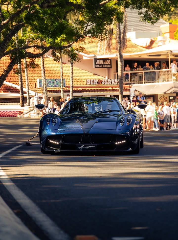 Blue exposed-carbon Pagani Huayra rolling down a Noosa street at golden hour with spectators lining the kerb.
