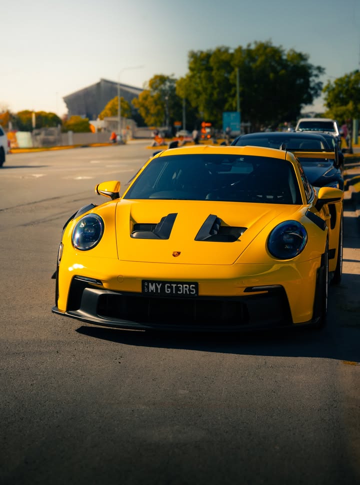 Yellow Porsche 911 GT3 RS head-on at a car meet, late afternoon sun raking across the louvred front guards.