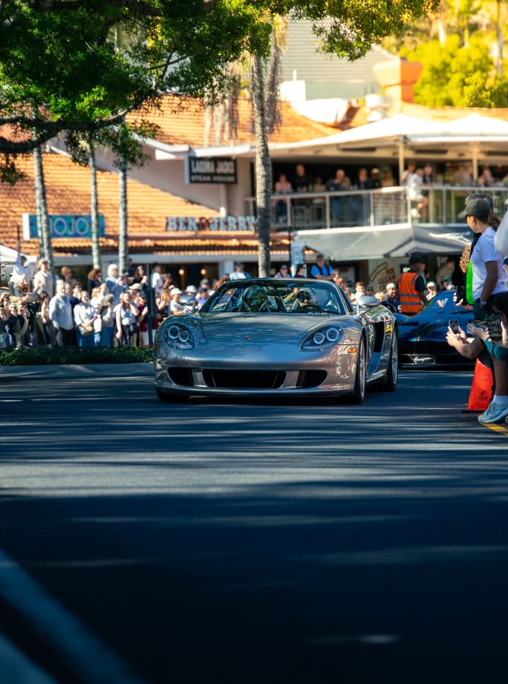 Silver Porsche Carrera GT rolling down a crowd-lined street at golden hour, a Pagani Huayra following just behind.